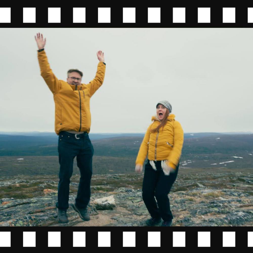 Mies ja nainen hyppivät innosta hihkuen tunturin huipulla. Heillä on yllään kirkkaan oranssit takit.