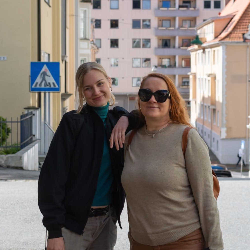 Vaaleahiuksinen nainen nojaa hieman lyhyemmän punahiuksisen naisen olkapäähän. Taustalla näkyy alaspäin viettävä kaupunkikatu.